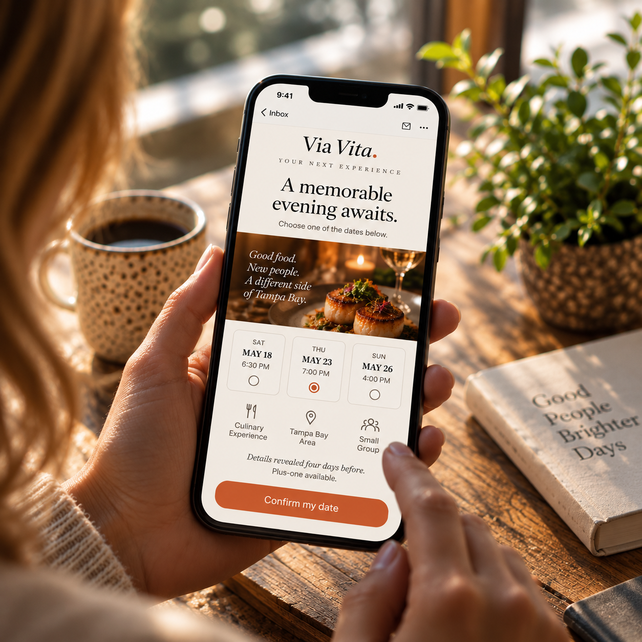 Phone in hand showing a Via Vita Club invitation with three date choices